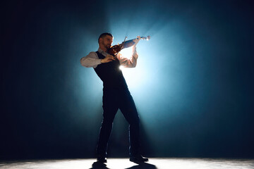 Classical musician performing on violin, playing melody on stage with spotlight from behind him...