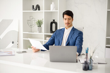 An industrious Hispanic businessman is engrossed in reviewing a paper document while managing tasks on his laptop, exemplifying the multitasking demands of current corporate roles