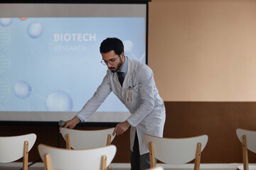 Medium full shot of young Muslim man doctor arranging chairs before start of medical presentation in conference room