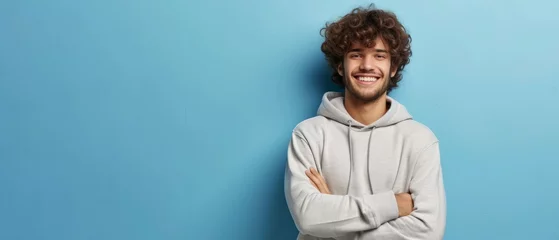 Foto op Plexiglas Portrait of happy man with curly hair smile toothily keeps arms down wears casual hoodie looks cheerful isolated over blue background being in good mood with copy space. Generative ai © Mickael