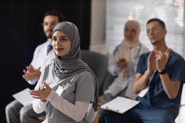 Medium shot of smiling Islamic woman wearing gray hijab applauding with audience to speaker during medical conference