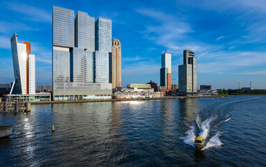 Obraz premium Rotterdam waterfront panorama from “Erasmus-Bridge“ over river Nieuwe Maas on a blue sky day in South Holland Netherlands. Tall office building skyline, water taxi speed boat near major sea port.