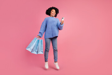Full length photo of lovely young lady hold shopping bags jump gadget wear trendy blue knitwear garment isolated on pink color background