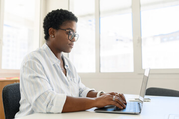 Confident serious young business woman, female freelancer, programmer, ceo corporate leader wearing smart casual shirt sitting inside and using laptop in office, typing