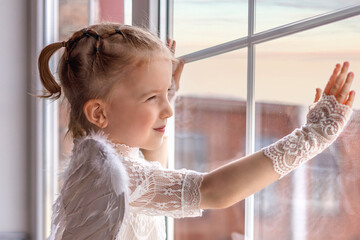 A little angel stands on the windowsill and looks out the window. Cute little angel girl looks and...