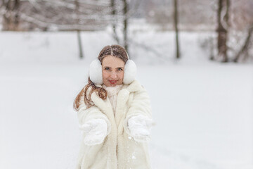 Fototapeta na wymiar Happy young woman in a white fur coat, mittens and fur headphones on a winter walk in the park