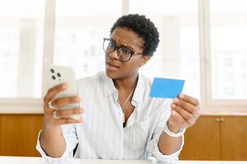 Obraz na płótnie Canvas Confused young african-american woman with short hair holding smartphone and credit card, has debt, female office employee or freelancer not received salary, checking account balance with sad face