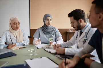 Experienced Muslim woman doctor in gray hijab giving speech during staff training while sitting at desk in meeting room