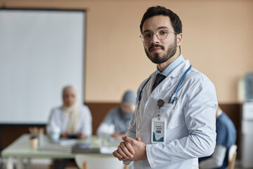 Confident young Muslim man doctor wearing eyeglasses and lab coat looking at camera and holding...