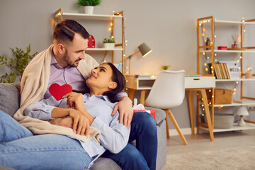 Obraz na płótnie Canvas Young cheerful smiling woman and man sitting on the couch at home, holding paper heart and looking tenderly at each other. Happy couple in love. Romance and Valentines day concept.