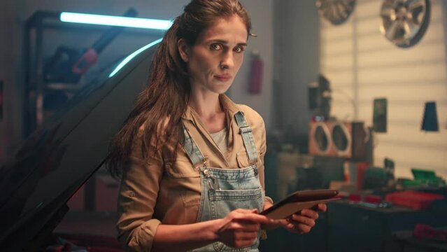 Portrait of beautiful woman standing in garage and looking at camera. Female auto mechanic orders tools, instruments for machine repair online using tablet. Service and maintenance of cars.
