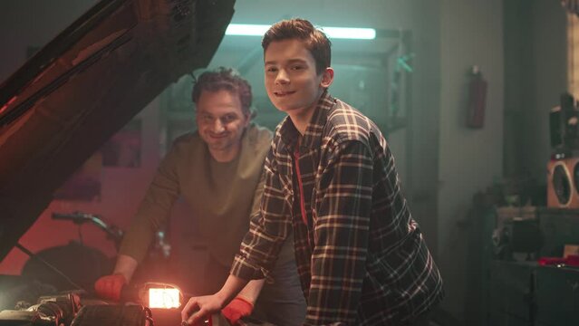 Background of workshop in dim light. Son helps father repair broken car engine in home garage. Maintenance service and small family business. Inspection of engine and checking motor oil level.