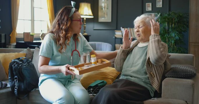 An old retired woman does not want to drink medicine she receives from the caregiver who takes care of her at home. Old female patient refusing to take medicine from nurse