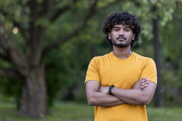 Portrait of a serious and confident Indian male athlete standing outside in a park in casual clothes, looking at the camera with his arms crossed on his chest