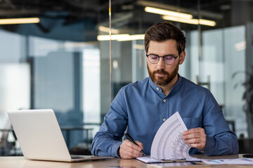 A young serious business man is sitting in the office at a desk with a laptop and is focused on...