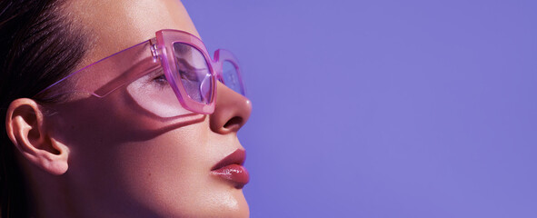 Profile portrait of a beautiful model in trendy square pink sunglasses with glossy make up posing against violet background.  Stylish glasses. Glance glamour picture. Summer Eye wear fashion .. - obrazy, fototapety, plakaty