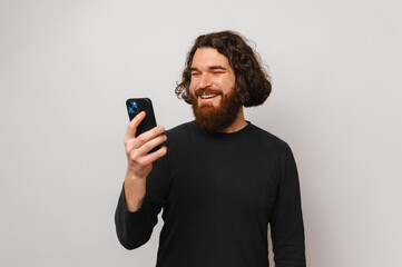 Wide smiling bearded man with long hair wearing black is using his smart phone. Studio shot over grey background.