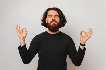 Handsome bearded man stands with eyes closed and holds fingers in zen gesture. Studio shot over...