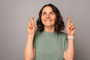 Cheerful middle aged woman is holding fingers crossed for a wish she made. Studio portrait over grey background.
