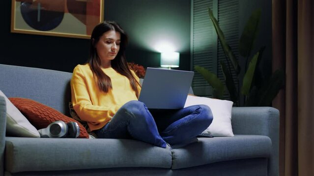 Portrait of cute young joyful woman working on computer, sitting on sofa at home, typing, chatting, communicates in social networks, checking emails on laptop. Leisure time activities.