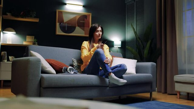 Young woman relaxing on couch at home enjoying television soap opera with happy ending. Young lady sitting on comfortable sofa in living-room and watching romantic series or reality show on TV.