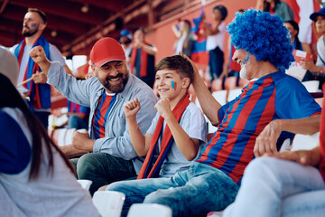 Happy kid cheering with his father and grandfather during sports game at stadium. - obrazy, fototapety, plakaty