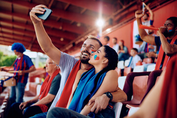 Young couple having fun while taking selfie during sports game at stadium.