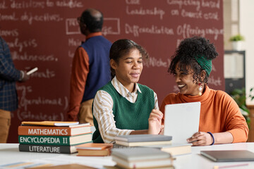 Portrait of two cheerful Black girls laughing while enjoying schoolwork in college classroom copy...