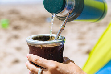 Woman serving hot water of a thermos in a Mate with yerba. Drinking Mate on the beach.