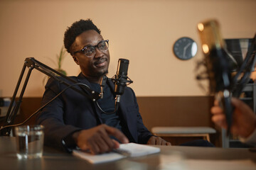 Medium shot of friendly Black man in eyeglasses and suit listening to unrecognizable podcast host question while sitting in studio - obrazy, fototapety, plakaty