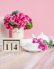 Wooden calendar with Valentine's Day for February 14 on the background of flowers and a cup of hot drink for breakfast