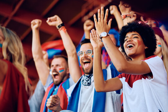 Happy black woman cheering with group of fans during sports match at stadium.