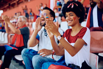 Happy black woman applauding while cheering for sports team during game at stadium.