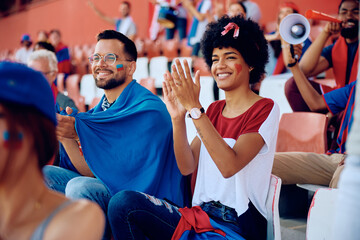 Happy black woman applauding while watching sports game at stadium.
