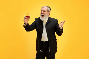Surprised mature man in kippah and formal black suit drinks alcohol drink against yellow background. Happy Purim. Festival, holiday, celebration Pesach or Passover, Judaism, religion concept. Ad - obrazy, fototapety, plakaty