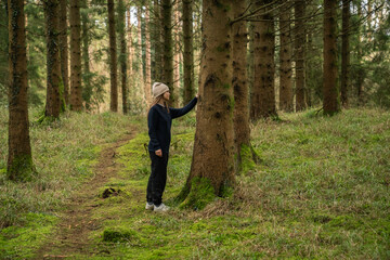 middle-aged white woman with brown hair, wearing a black sweater and a beige hat, in an Irish winter forest.