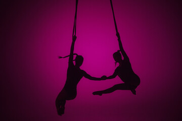 Silhouette of two female acrobats isolated on pink neon background. Girls aerial dancers performing flying element on ropes.