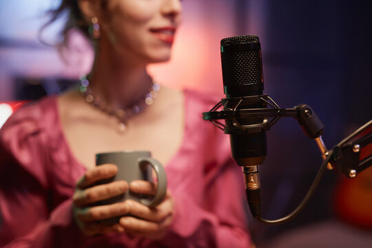 Close up shot of black condenser microphone in holder and unrecognizable female podcast host with cup in studio