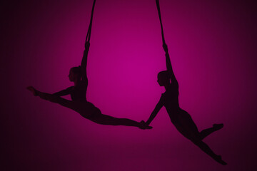 Silhouette of two female acrobats isolated on pink neon background. Girls aerial dancers performing flying element on ropes.