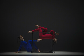 Portrait of two female acrobats isolated on black background. Girls aerial dancers in blue red suits showing element on a cube.