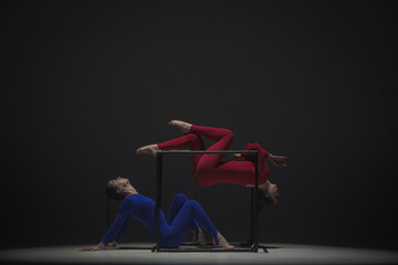Portrait of two female acrobats isolated on black background. Girls aerial dancers in blue red suits showing element on a cube.