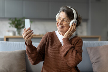 Modern senior woman in headphones engages in vibrant moment, capturing selfie on cellphone or connecting through video call, sitting on couch at home