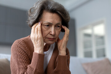 Sad, depressed senior woman sitting on sofa, touching her temples in visible distress, suffer from headache, challenges of physical and emotional well-being in aging process