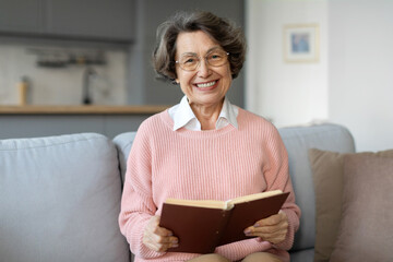 Happy retired senior woman reading book, sitting on couch and smiling at camera, enjoying free time...