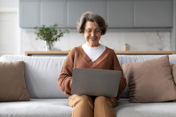 Cheerful senior woman sitting on her home sofa, radiating joy as she engages with laptop, living...