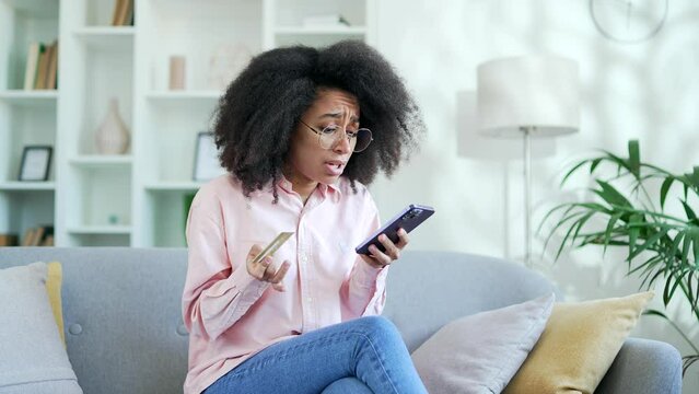 Shocked upset young african american female discovered fraud entering credit card number on smartphone sitting on sofa at home. Frustrated black woman saw that money had been stolen from her account