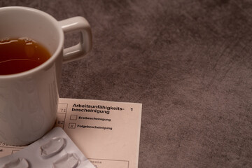 Eine Tasse Tee neben Medikamenten und einer Arbeitsunfähigkeitsbescheinigung auf grauem...