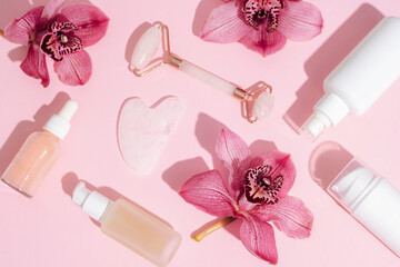 Facial cosmetic roller and gua sha scraper with pink orchid flowers and serum bottles. Sharp shadows. Home spa concept. Top view, flat lay - obrazy, fototapety, plakaty