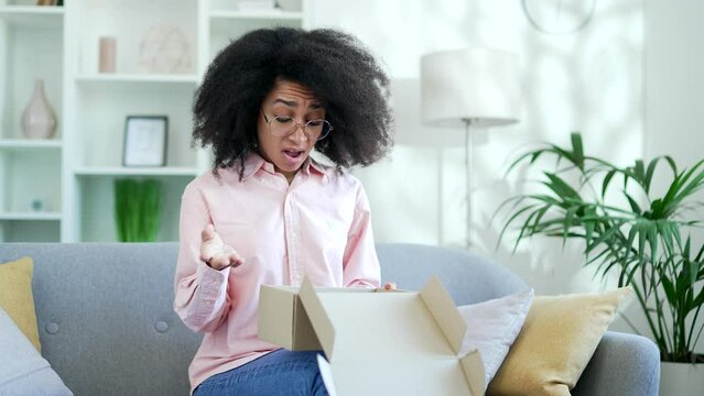 Unhappy african american female received a parcel with wrong product sitting on sofa in living room at home. Black woman is dissatisfied with shopping in an online store. Service issue or damaged item