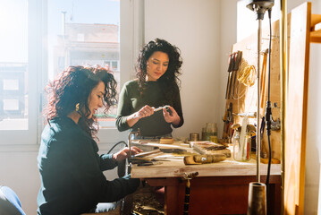 Twin sisters measuring and cutting jewelry in workbench on sunny day - obrazy, fototapety, plakaty
